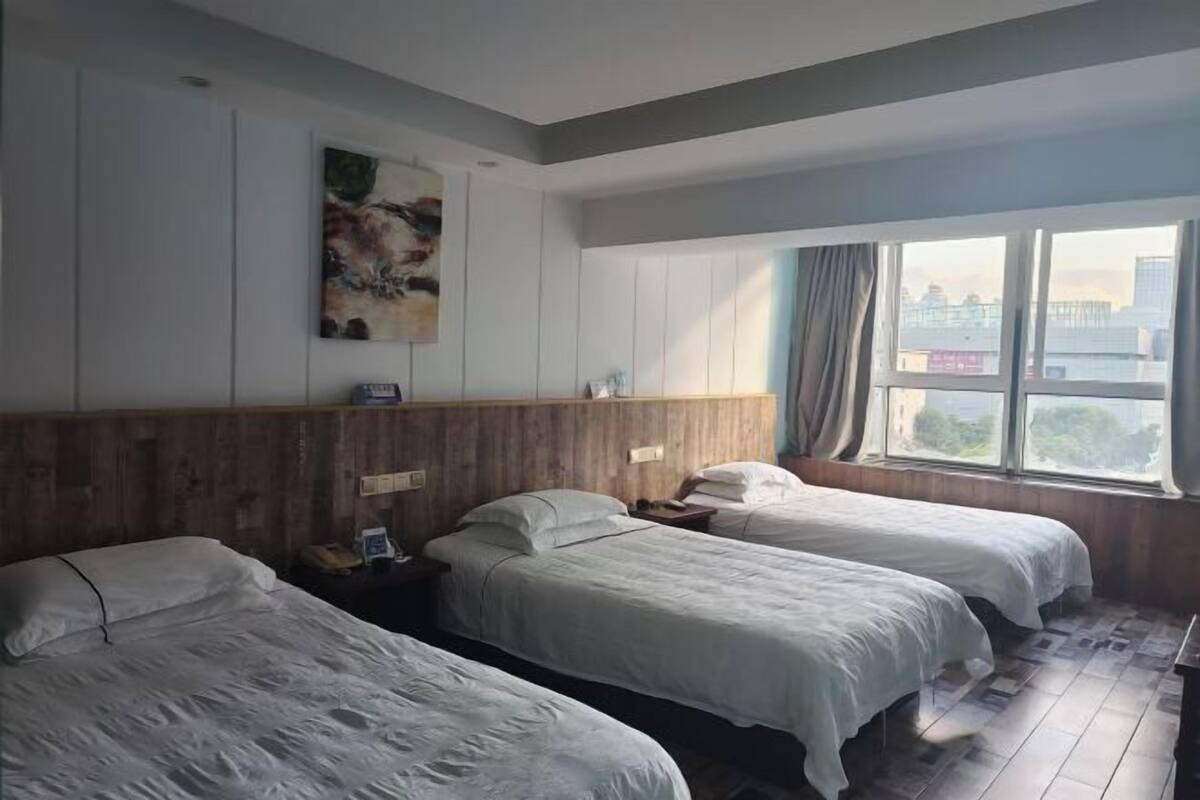 A hotel room with three beds, a wooden headboard, a painting, a window with a view of the city, and a nightstand with a lamp.