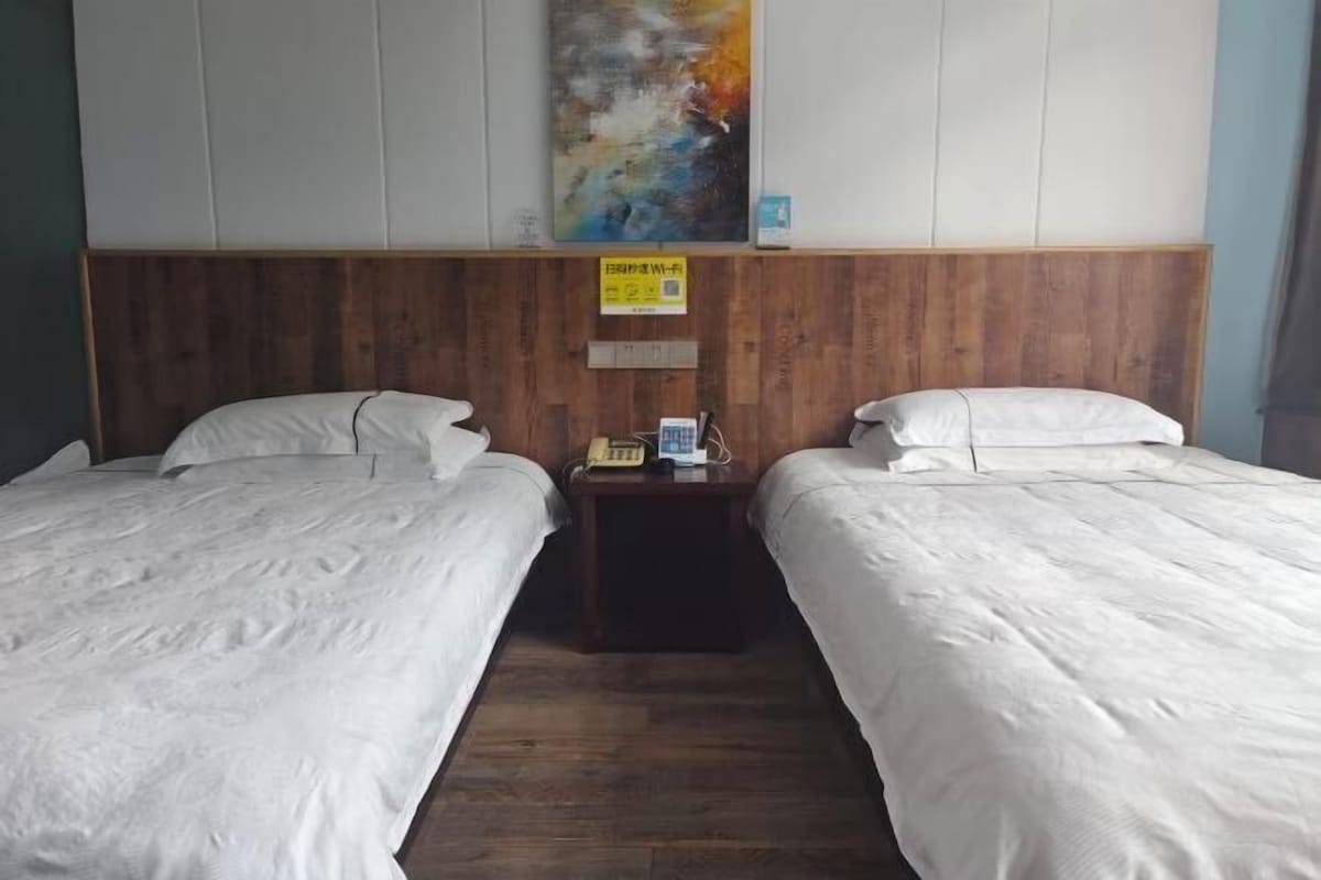 Une chambre d’hôtel avec deux lits simples, une tête de lit en bois, une petite table de chevet avec un téléphone et un panneau, et une peinture abstraite aux couleurs vives accrochée au mur.