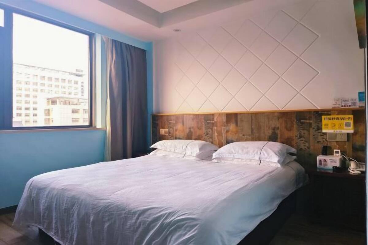 A hotel room with a bed, a window with curtains, a wooden headboard, and a wall-mounted Wi-Fi sign.