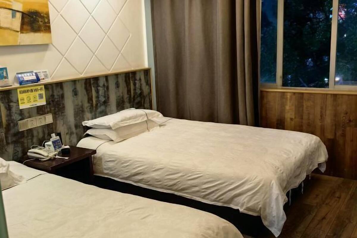A hotel room with two beds, a wooden headboard, a nightstand with a phone, and a window with a view of trees.