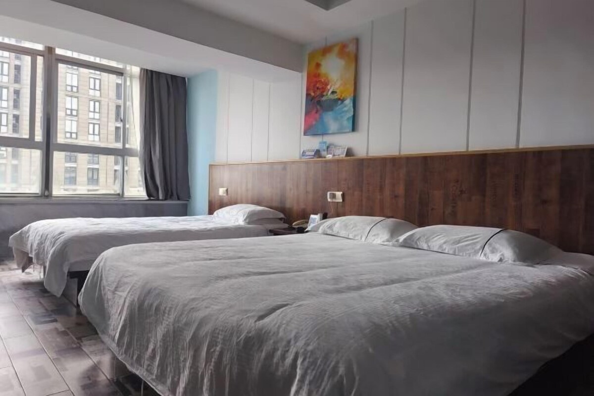 Une chambre d’hôtel avec deux lits, une grande fenêtre avec des rideaux, une tête de lit en bois et une peinture abstraite aux couleurs vives accrochée au mur.