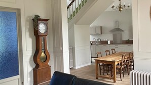 A living room with a black leather sofa, a dining area with a wooden table and chairs, a kitchen with white cabinets, and a staircase leading to an upper floor. - Le Clos de Lignières : Chez Marguerite (Lignières-Châtelain)