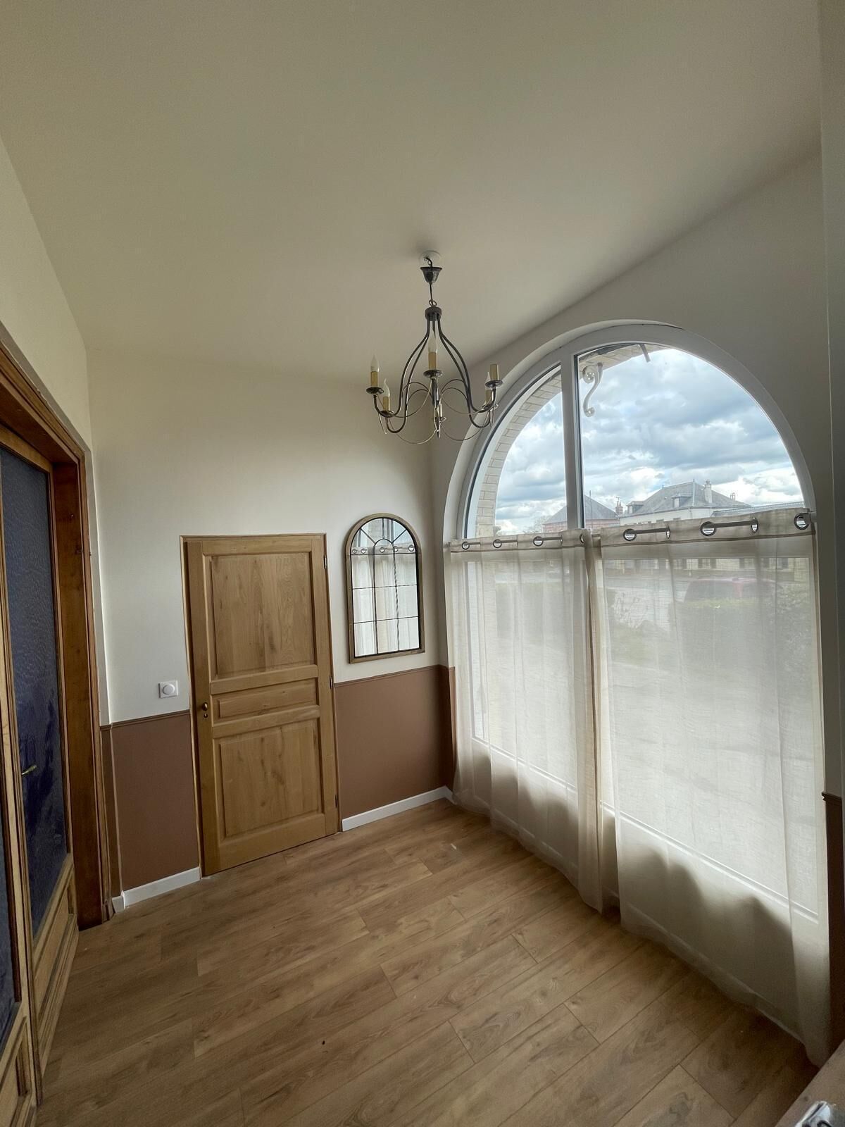 Ein Zimmer mit Holzboden, einem großen Bogenfenster mit Blick auf Gebäude, einer Holztür und einer Chandelüster.