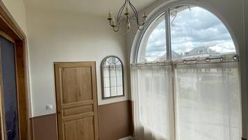 Ein Zimmer mit Holzboden, einem großen Bogenfenster mit Blick auf Gebäude, einer Holztür und einer Chandelüster.