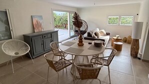 Un grand espace dédié à la salle à manger, avec une table ronde et des chaises en osier, un buffet gris et une vue sur la verdure à l’extérieur.