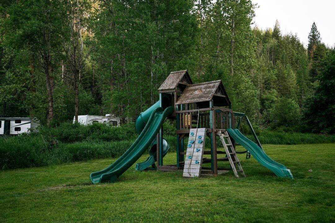 A wooden play structure with two slides in a grassy area surrounded by trees.