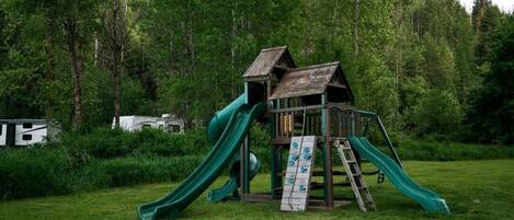 A wooden play structure with two slides in a grassy area surrounded by trees.