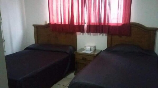 A room with two single beds, a wooden headboard, a nightstand with a lamp, and a window with red curtains.