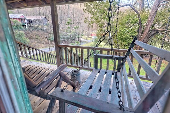 A wooden swing chair on a porch with a view of a house and trees.