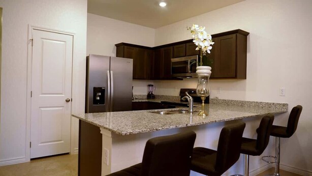 A kitchen with dark wooden cabinets, a granite countertop, a stainless steel refrigerator, a built-in microwave, and a vase with white flowers. - Peaceful Stay Near Fort Cavazos (Copperas Cove)
