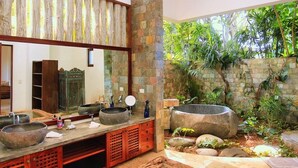 Un baño con un espejo grande, dos lavamanos de piedra y un lavabo de piedra. Hay un mueble de madera con cajones y una repisa. El baño tiene una pared de piedra y vistas a la vegetación exterior.