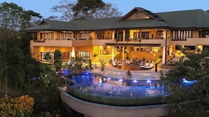 Un hotel grande de varios pisos con una zona de piscina, mobiliario exterior y una entrada bien iluminada.