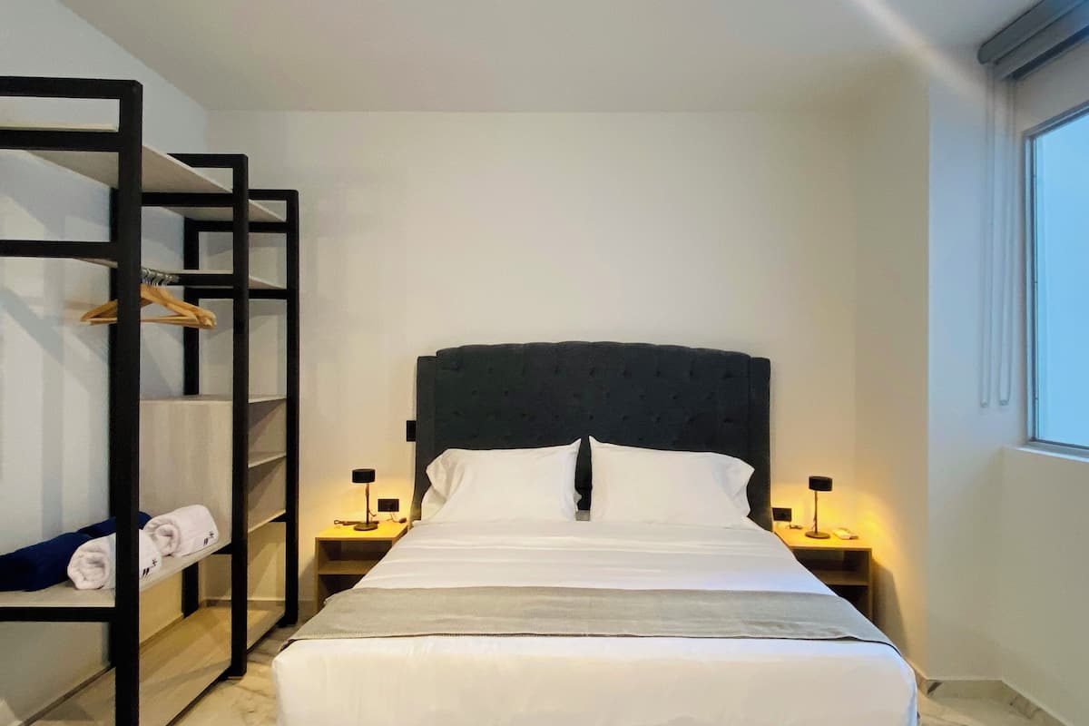 Una habitación de hotel con cama, dos mesitas de noche, un estante en la pared con toallas y una ventana.