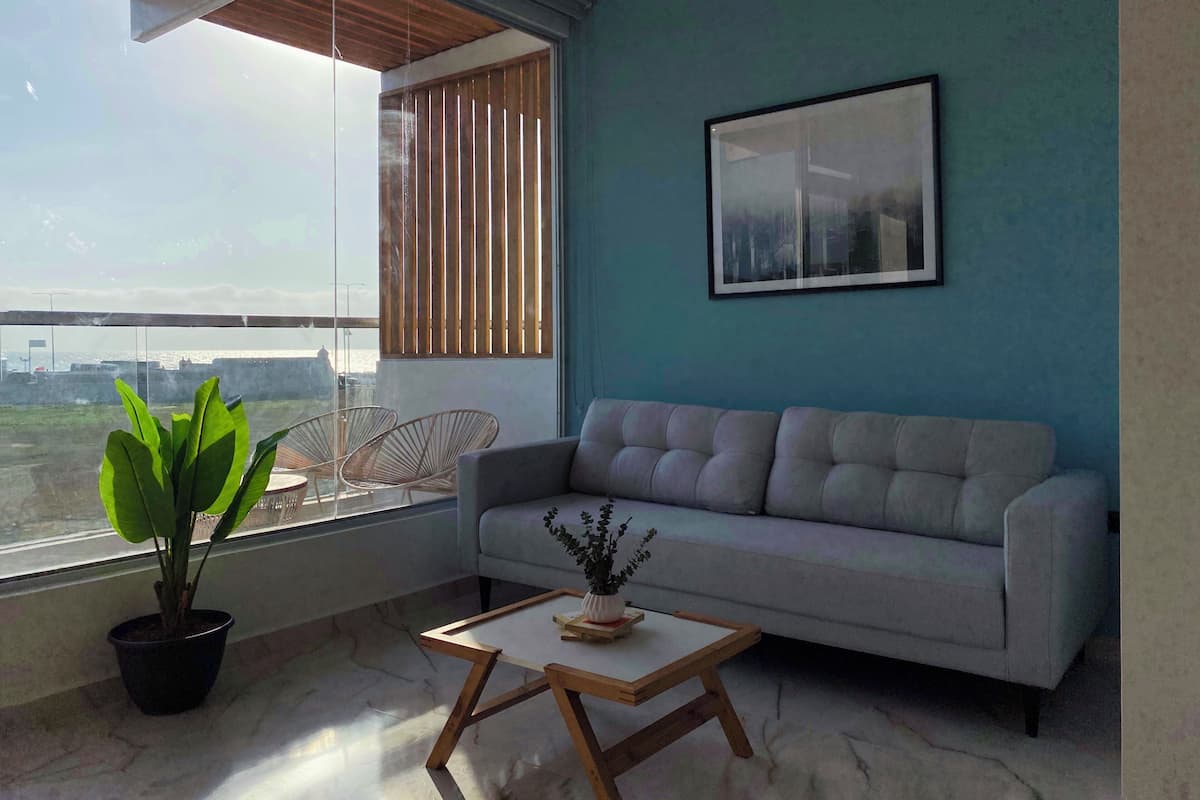 Una sala moderna con un sofá gris, una mesa de centro de madera y una planta en maceta junto a un ventanal con persianas.