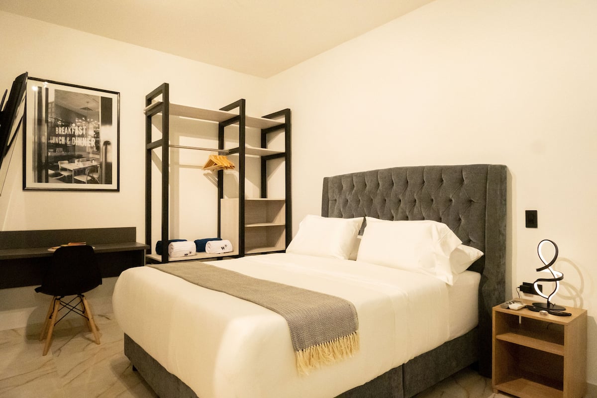 Habitación de hotel ordenada con una cama grande, un escritorio y una silla. Hay un cuadro en la pared y una pequeña estantería con una lámpara.