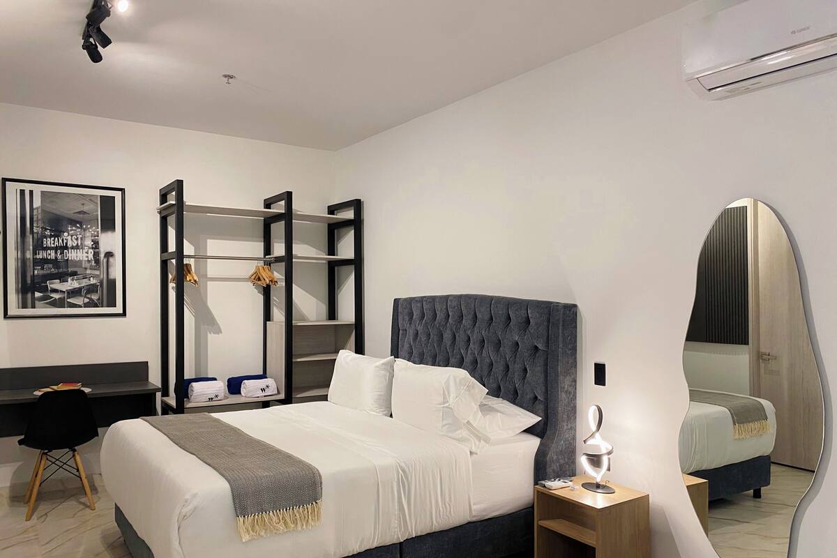 Una habitación de hotel moderna con una cama grande, una mesita de noche con lámpara, un armario, un escritorio y un espejo.