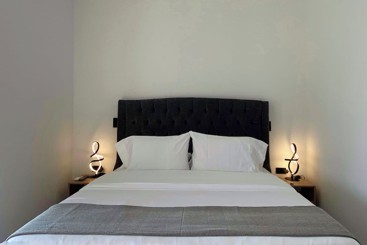 Una cama bien hecha con un cabecero oscuro, ropa de cama blanca y una manta gris. Hay dos mesitas de noche con lámparas a cada lado de la cama.