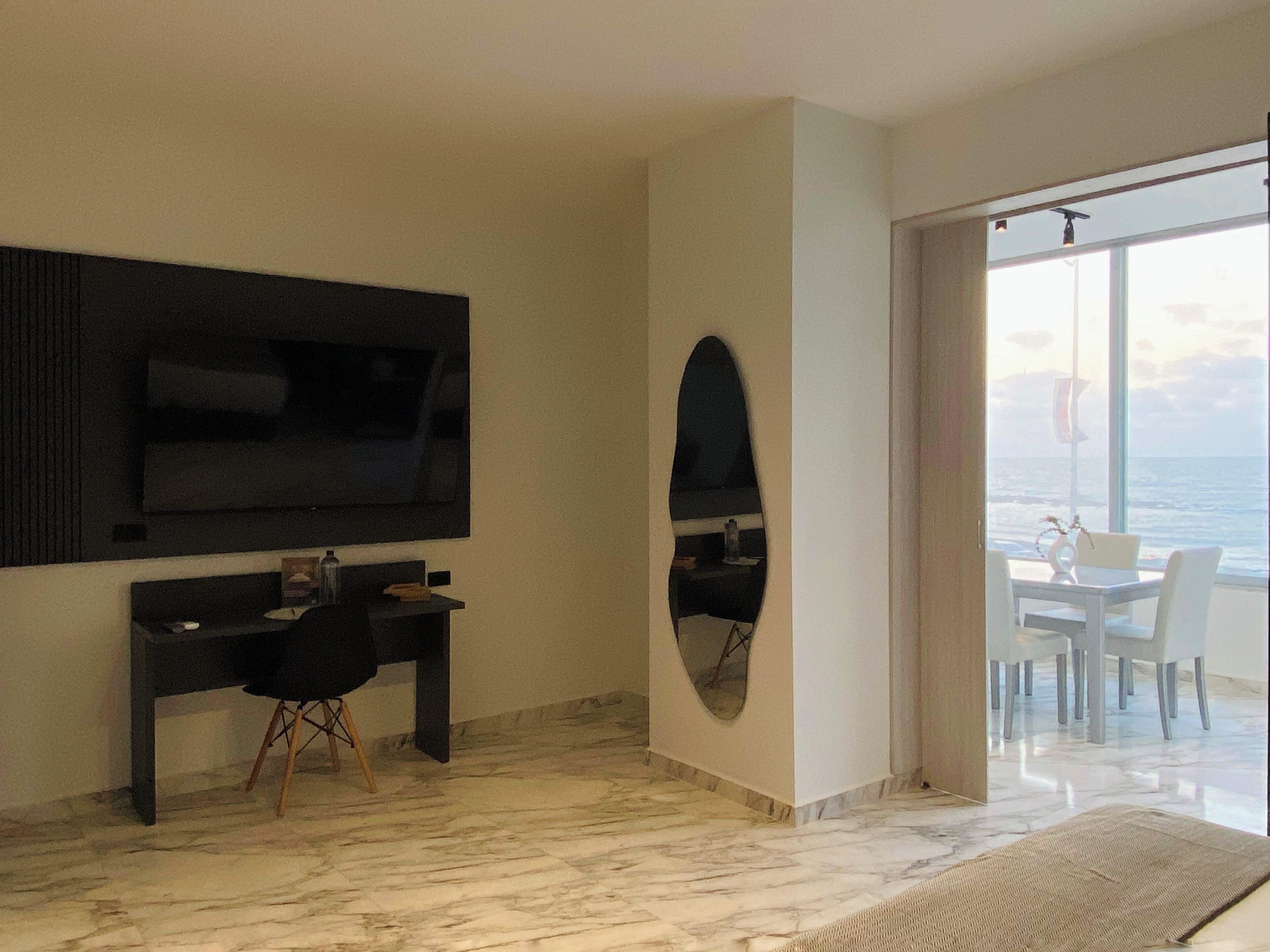 Habitación de hotel moderna con televisor grande, escritorio y zona de comedor con vista al mar.