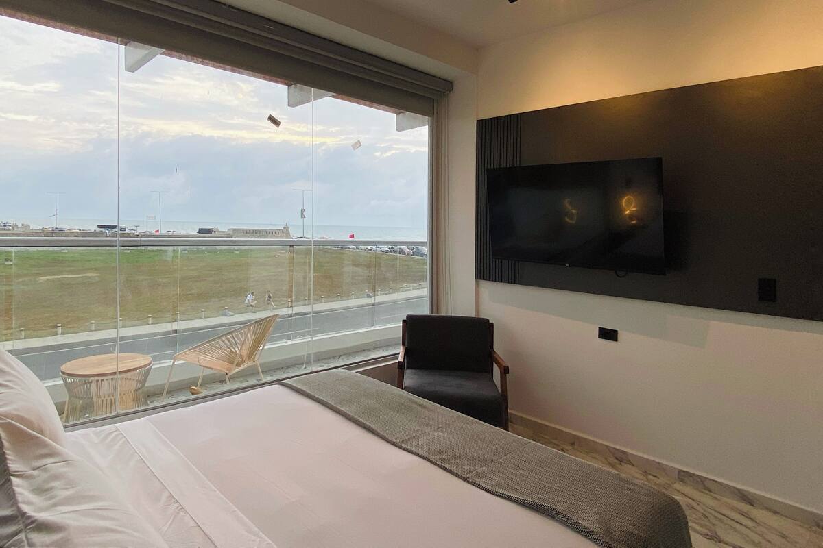 Habitación de hotel moderna con una cama grande, un televisor montado en la pared y vistas al aeropuerto a través de amplios ventanales.