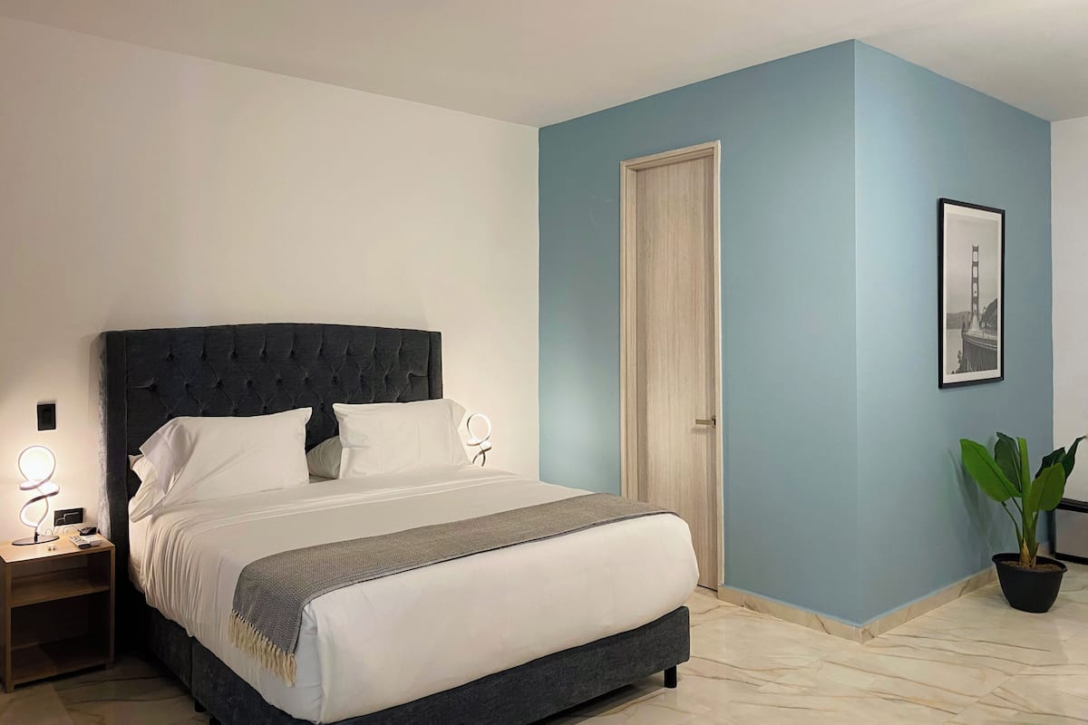Un dormitorio con cama de cabecera oscura, ropa de cama blanca, una mesita de noche con lámpara, una puerta y un cuadro en la pared.