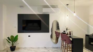 A modern living room with a large flat-screen TV mounted on the wall, a potted plant, a marble floor, a dining area with a round table and chairs, and pendant lighting.