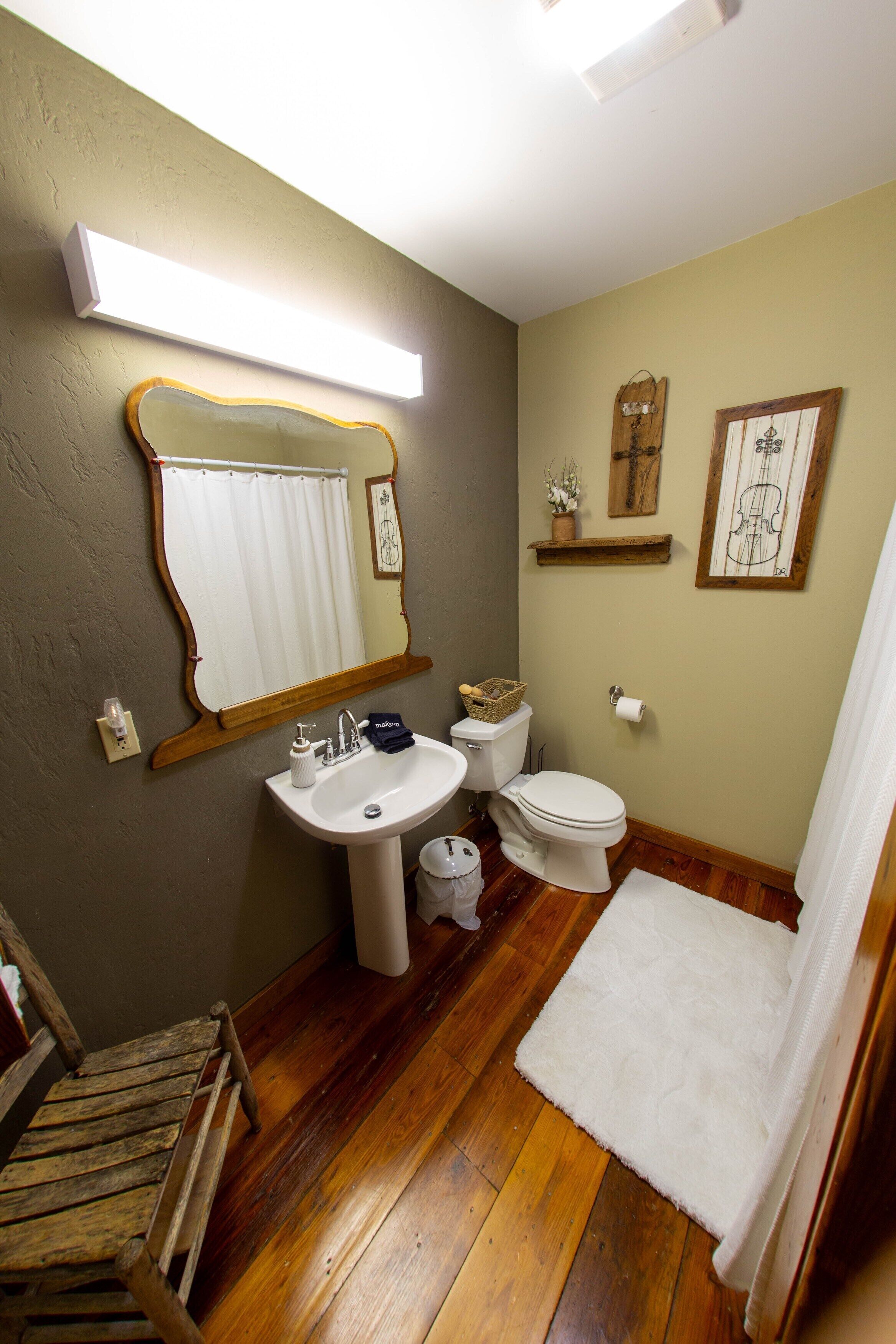 A bathroom with a wooden floor, a white sink, a toilet, a mirror, and a shower curtain.