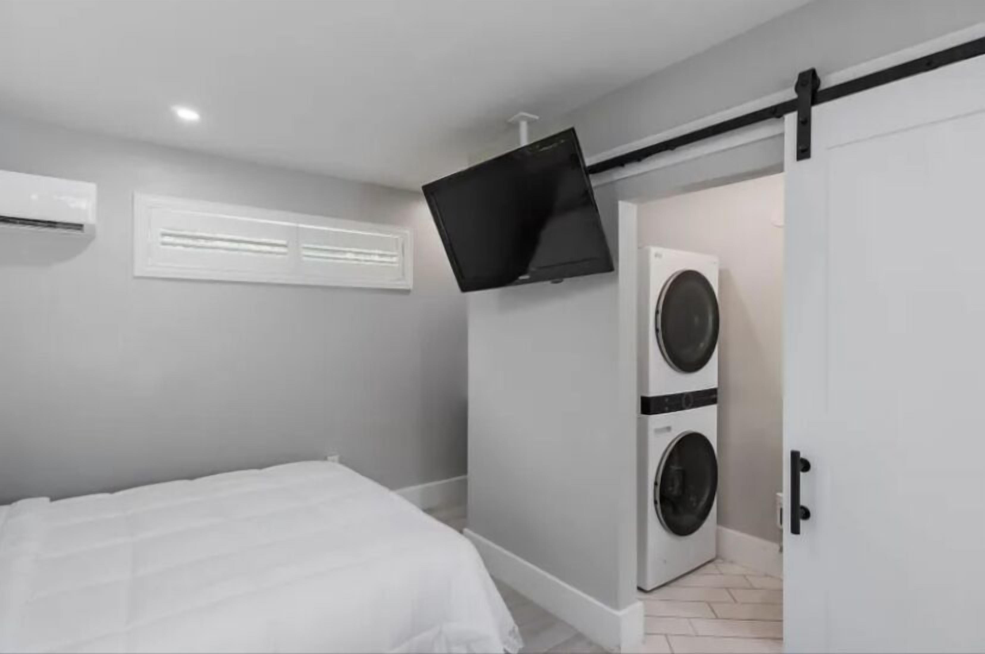 A compact room with a bed, a washing machine, and a television mounted on the wall.