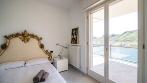A bedroom with a large bed, a nightstand, a radiator, and a view of a body of water through a sliding glass door.