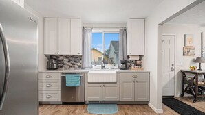 A modern kitchen with a stainless steel refrigerator, grey cabinets, a window with checkered curtains, a sink, and a coffee maker.