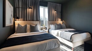A hotel room with two beds, a large window with curtains, a wall-mounted lamp, and a framed picture.
