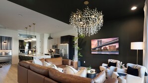 A modern living room with a brown leather sofa, white armchairs, and a chandelier. There is a kitchen in the background, a dining area, and a large window with curtains.