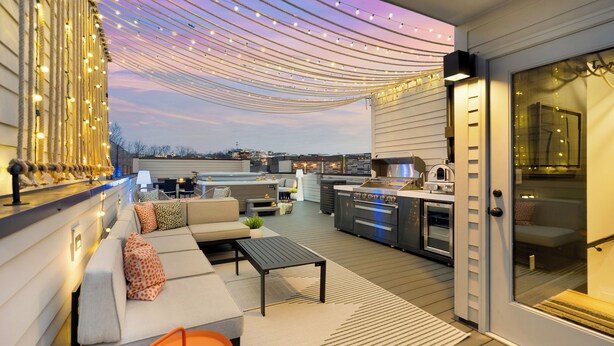 An outdoor deck with a sofa, table, and grill area, featuring string lights and a view of the city.