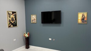 Een woonkamer met een flatscreen-televisie aan een donkerblauwe muur, grijze banken, een plafondventilator en twee kunstwerken met bloemen in lijsten.