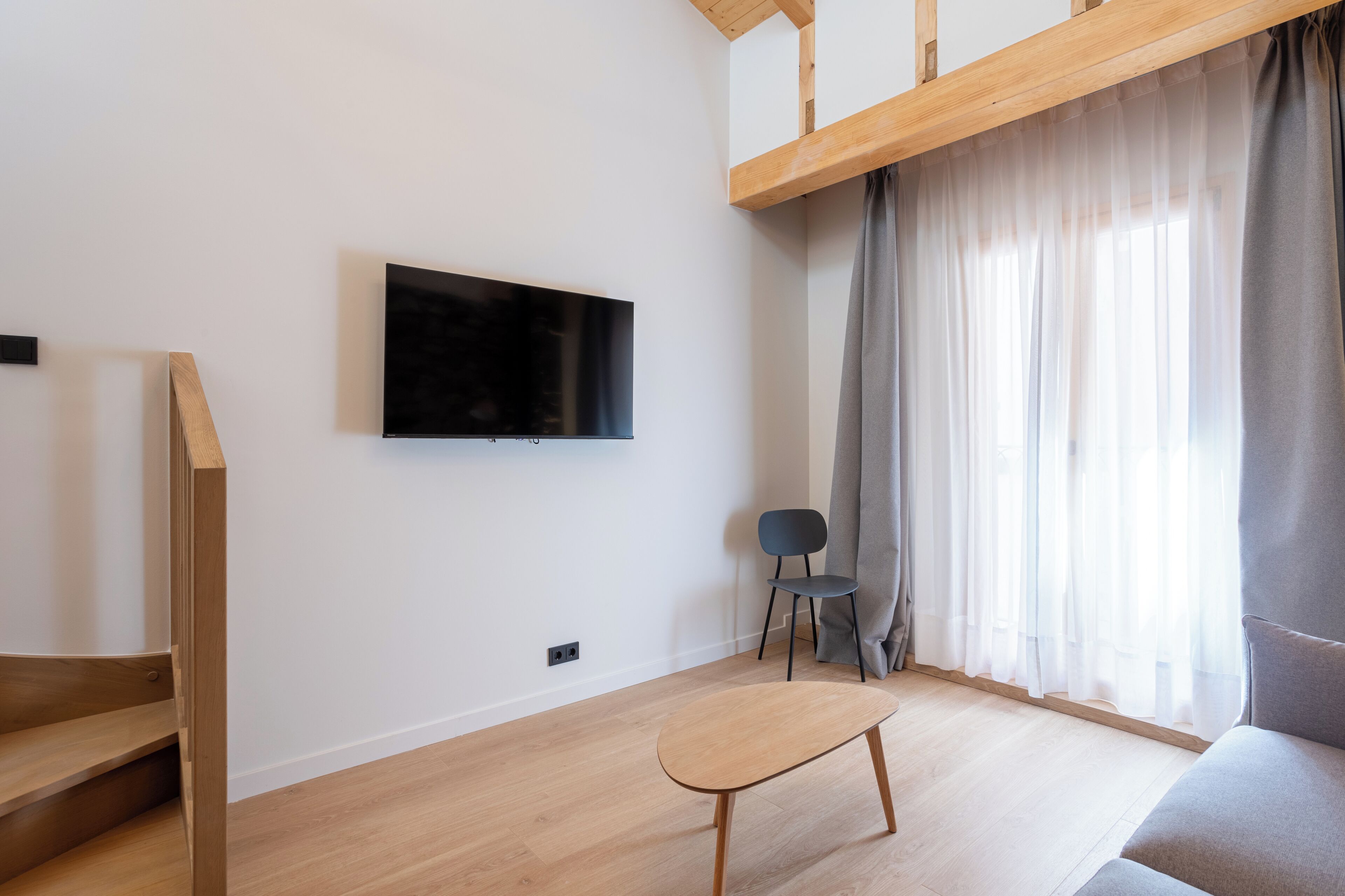 Una sala de estar moderna con una escalera de madera, un televisor de pantalla plana montado en la pared, un sofá gris, una mesita y una silla.