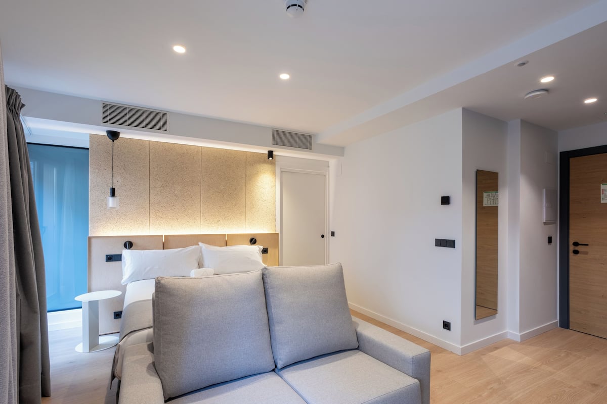 A modern hotel room with a sofa, a small table, a bed with white linens, a wall-mounted TV, and a wooden door.