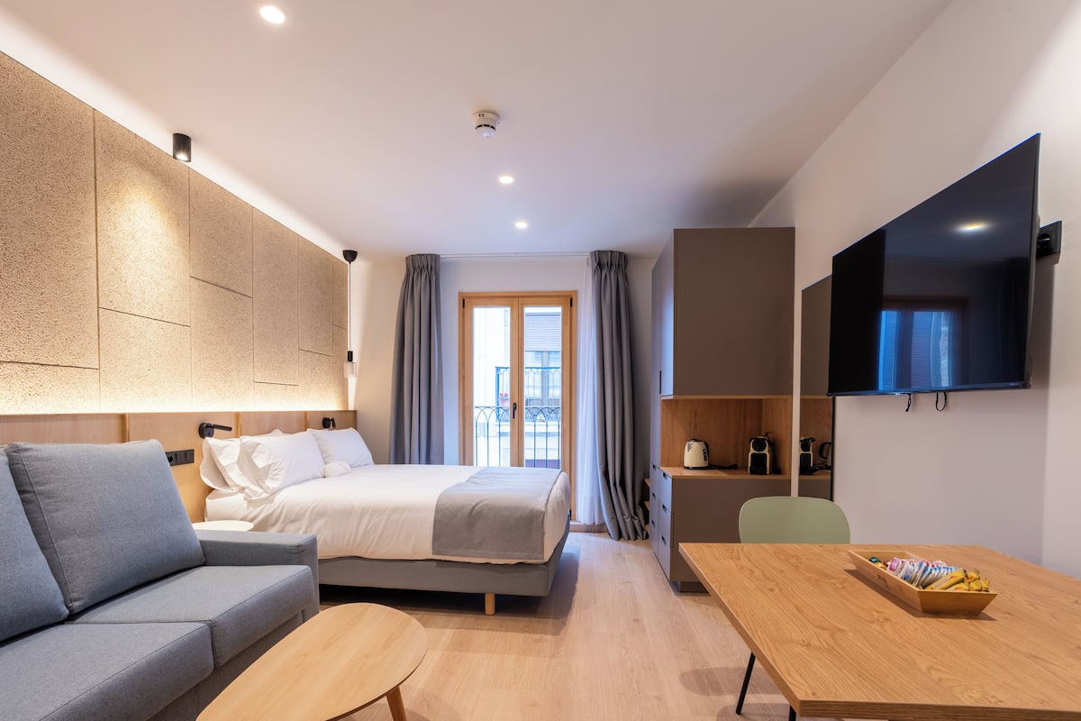 A modern hotel room with a bed, sofa, TV, and a dining area.