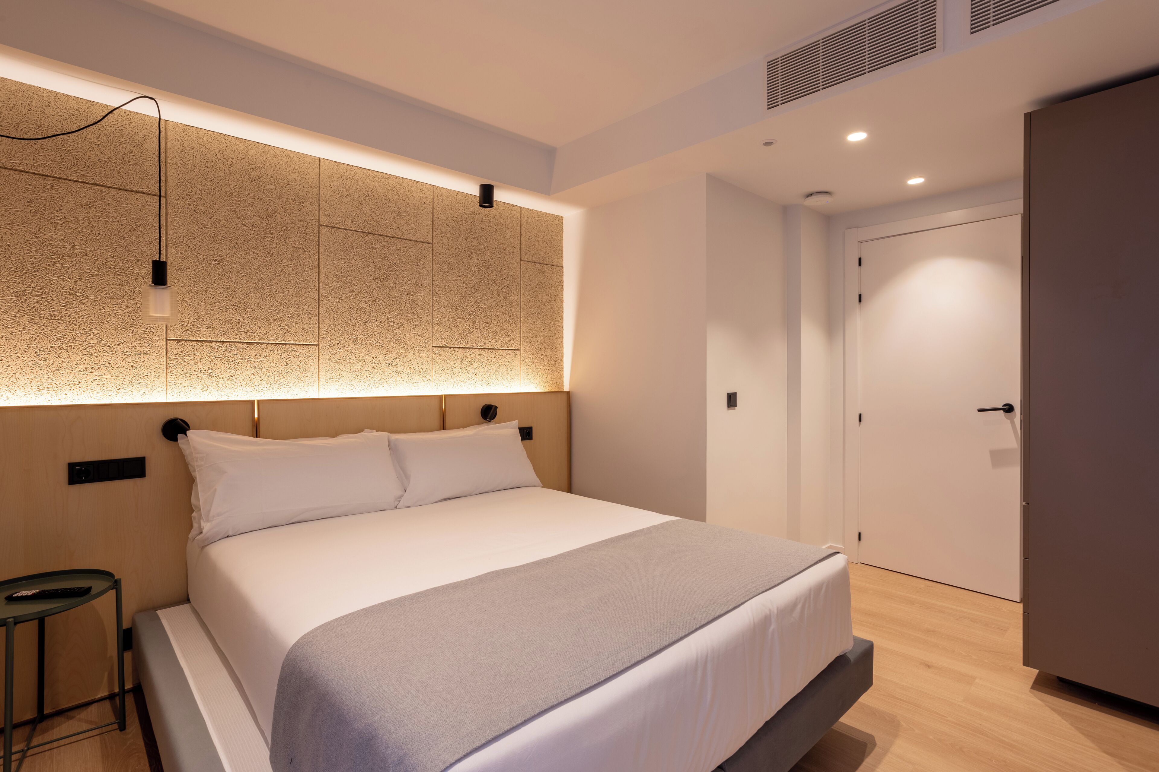 A modern hotel room with a large bed, a bedside table, a wall-mounted light, and a door.
