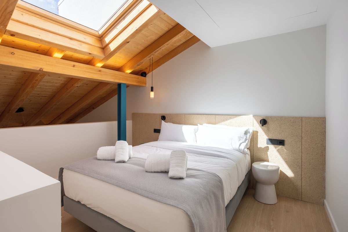 A modern bedroom with a skylight, a bed with white linens, a bedside table, and a toilet.