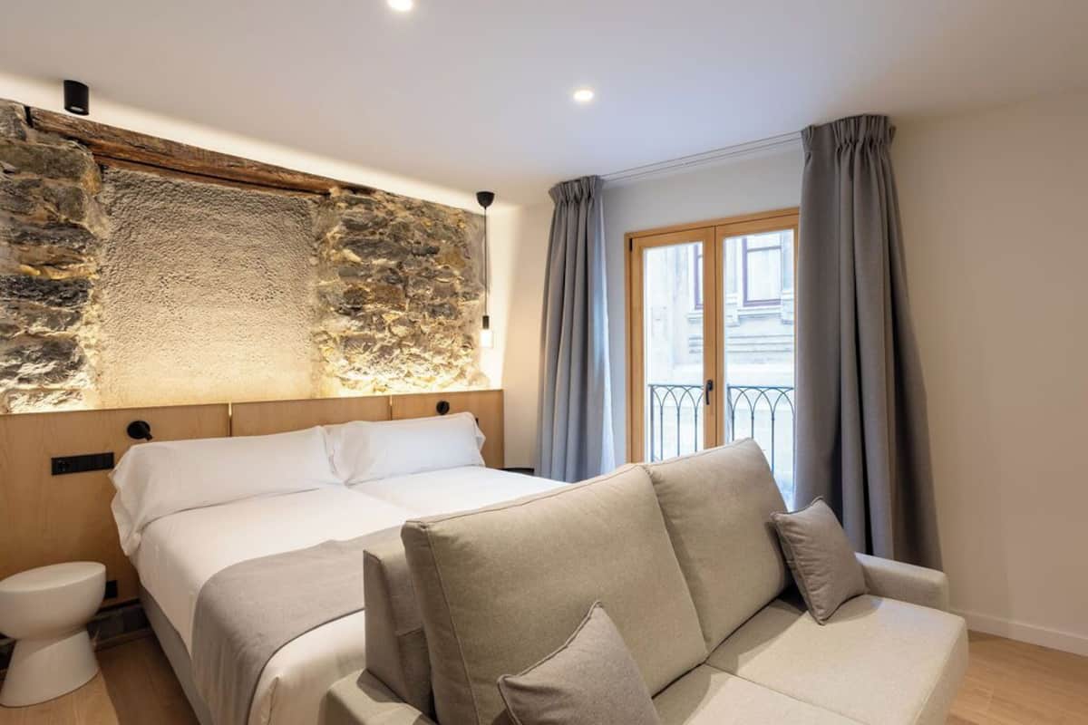 A bedroom with a stone accent wall, a double bed, a sofa, and a window with curtains.
