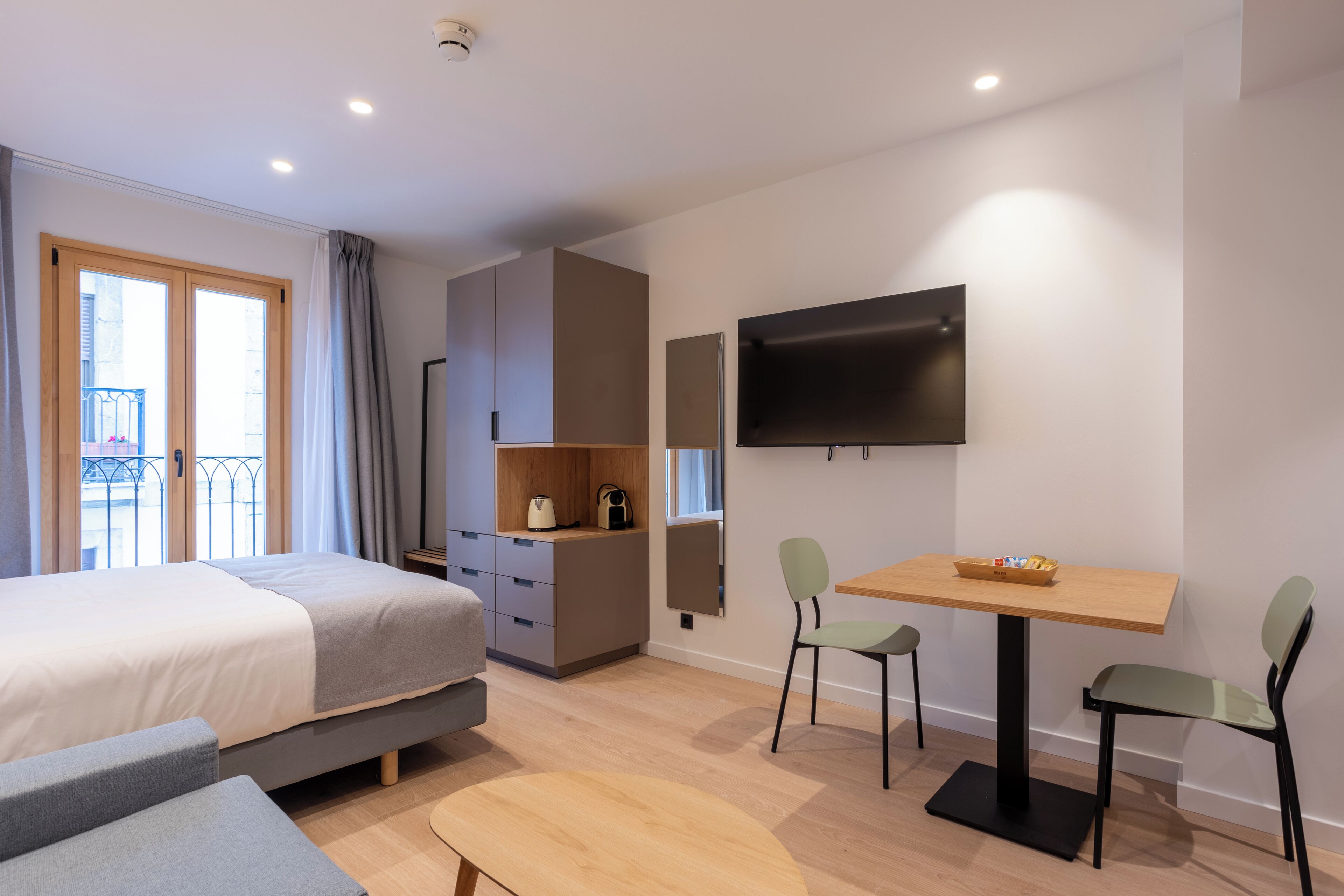 Une chambre d’hôtel moderne équipée d’un lit, d’un bureau, de deux chaises, d’une télévision et d’un balcon avec une rambarde.
