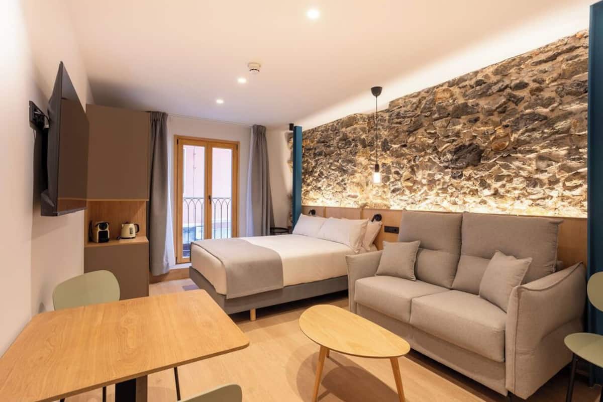 A modern hotel room with a stone accent wall, a bed, a sofa, a dining table, and a TV.