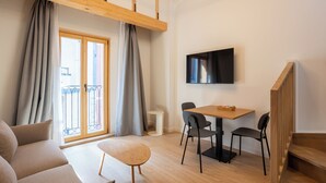 A modern living room with a sofa, a small table, a dining area with chairs, a TV mounted on the wall, and a balcony with curtains. - Be10 Suites (Bermeo)