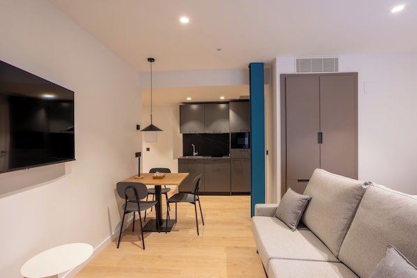 A modern living room with a sofa, a dining table with chairs, a TV mounted on the wall, and a kitchen area with cabinets and a microwave. - Be10 Suites (Bermeo)