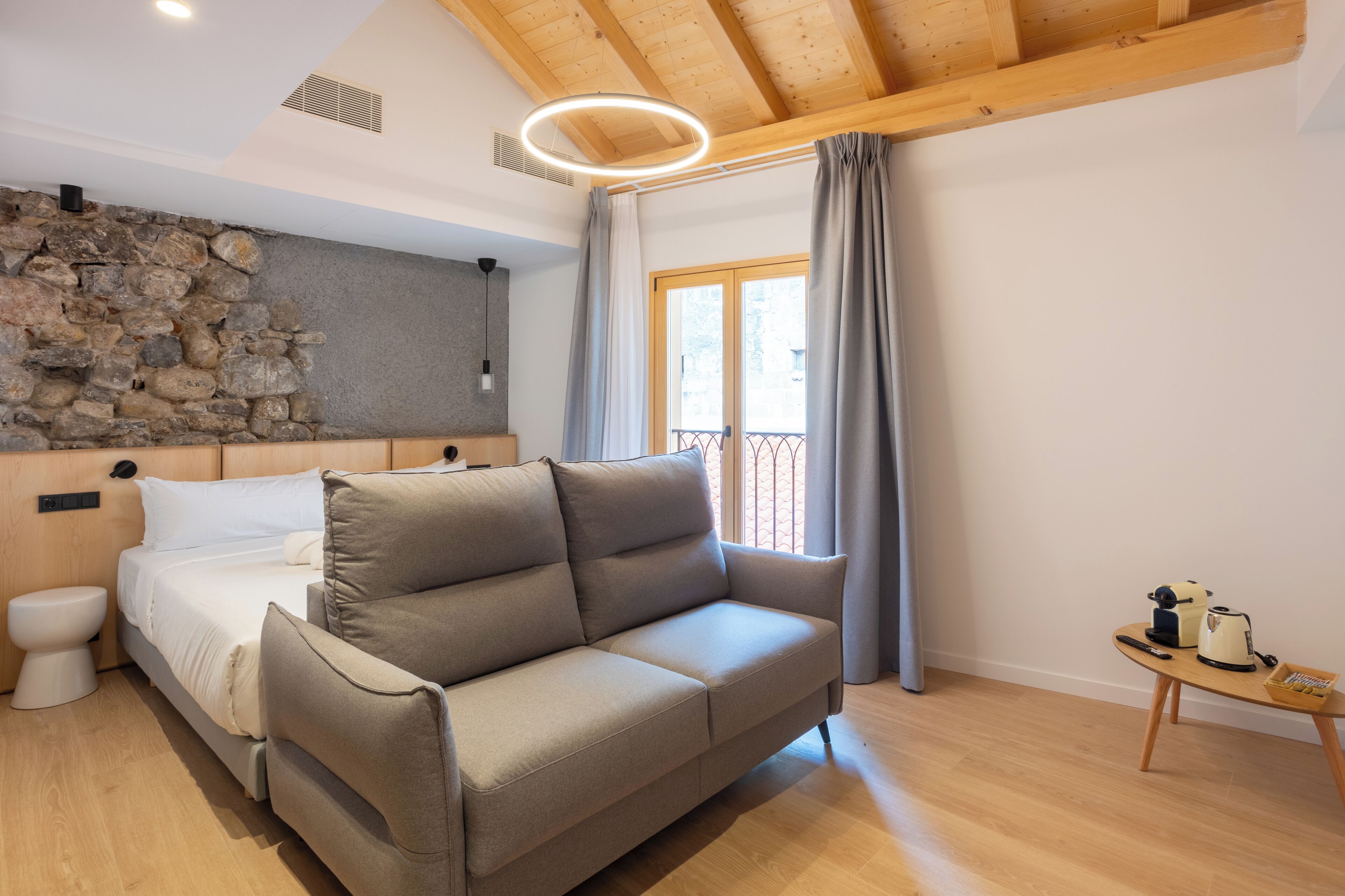 Un dormitorio moderno con pared de piedra, techo de madera, sofá gris, mesita de noche con hervidor y ventana con cortinas.