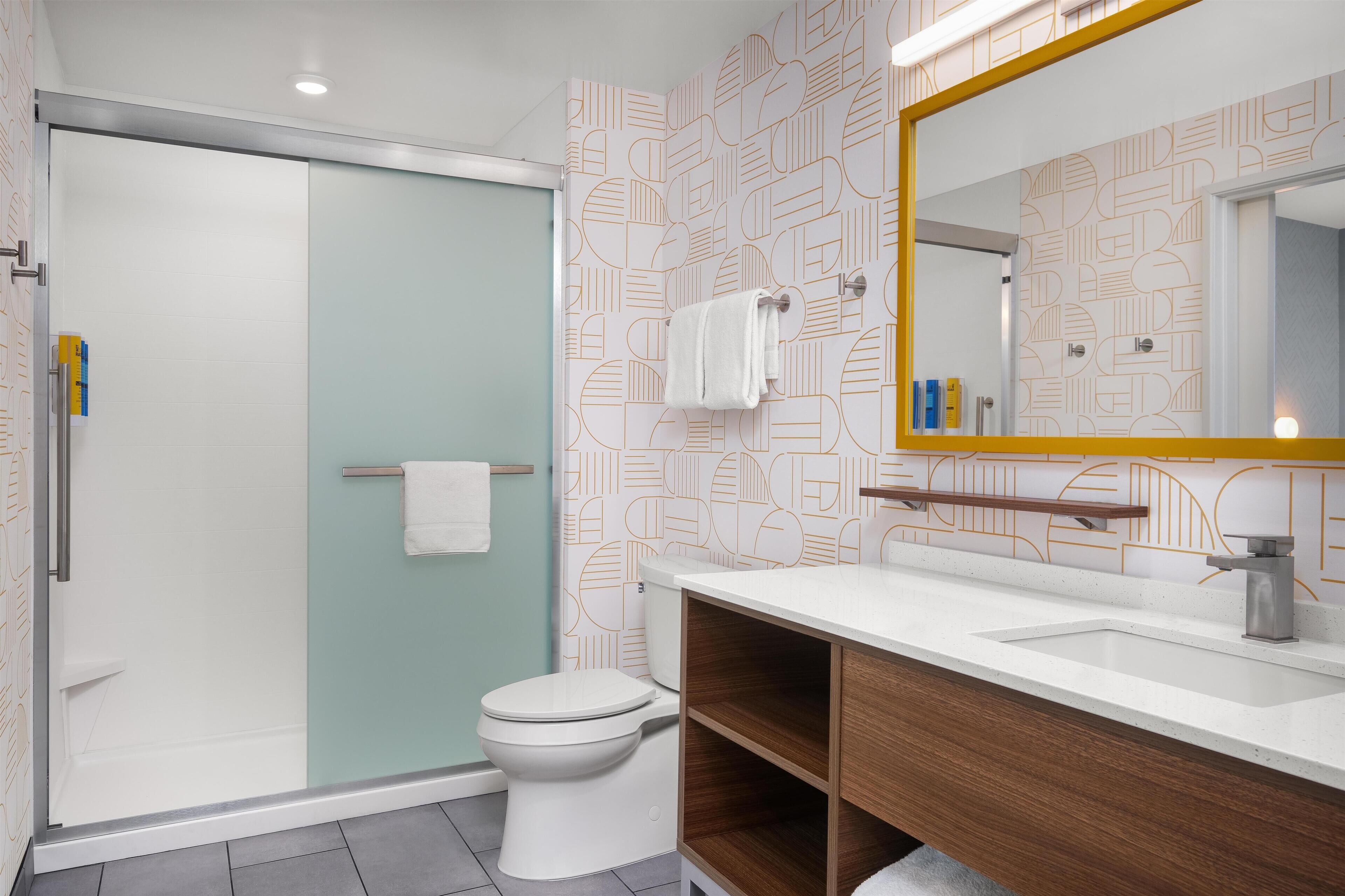 A modern bathroom with a glass shower enclosure, a white toilet, a wooden vanity with a mirror, and patterned wallpaper.