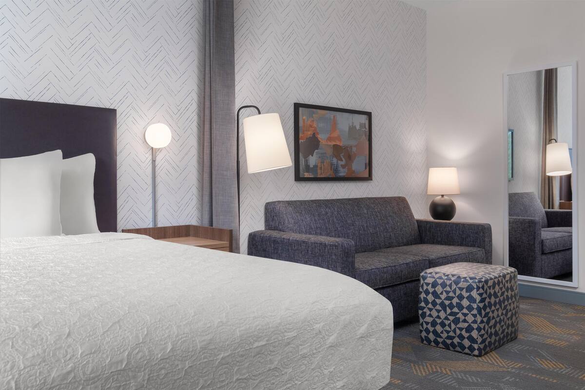 A hotel room with a large bed, a sofa, a small ottoman, a painting, and a mirror.