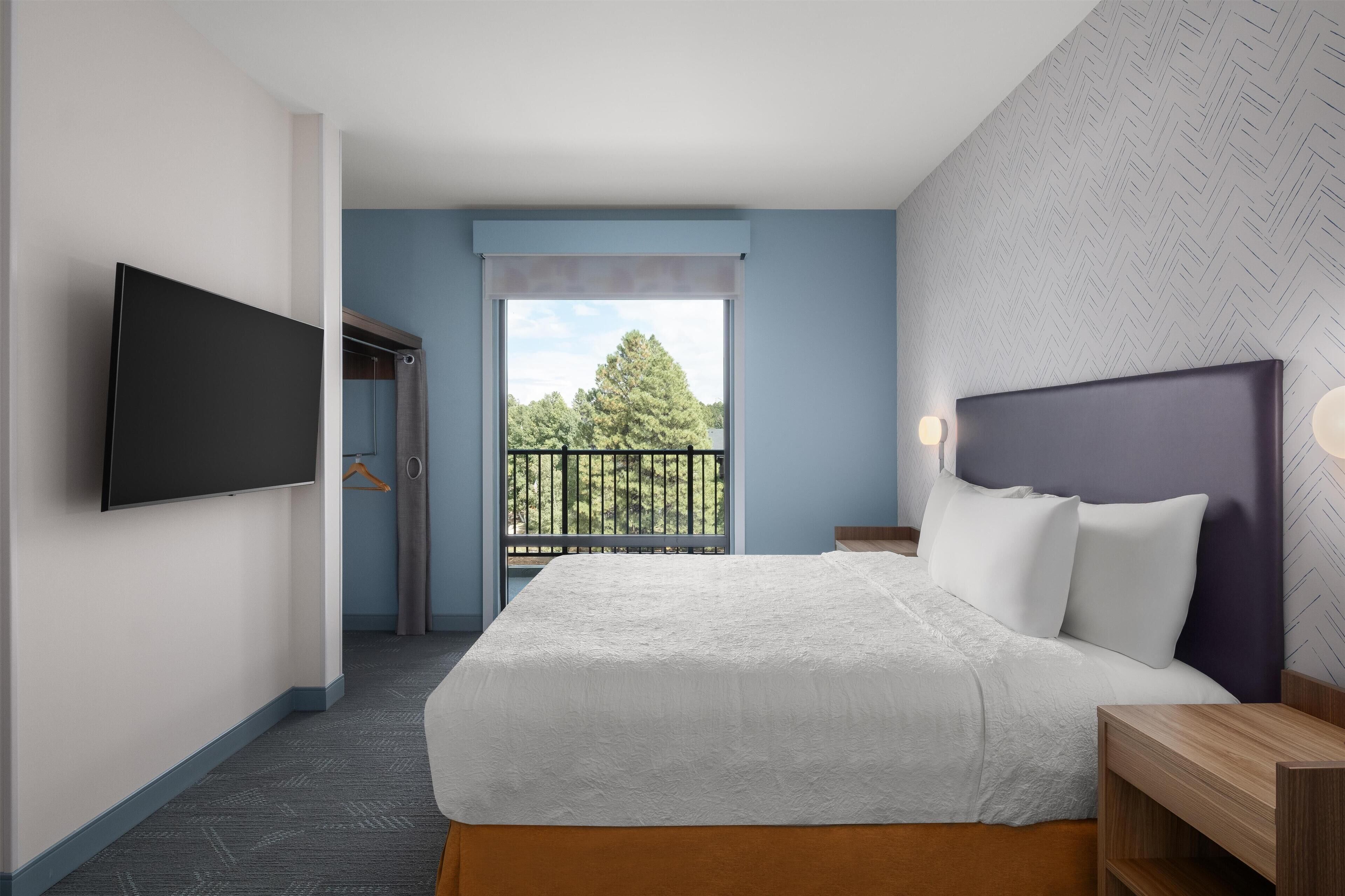 A hotel room with a bed, a TV, a balcony with a view of trees, and a patterned wall.