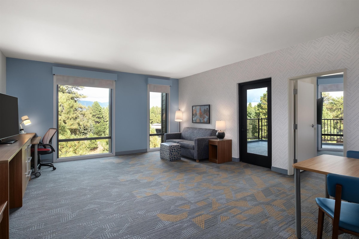 A hotel room with a desk, chair, sofa, and a balcony with a view of trees and mountains.