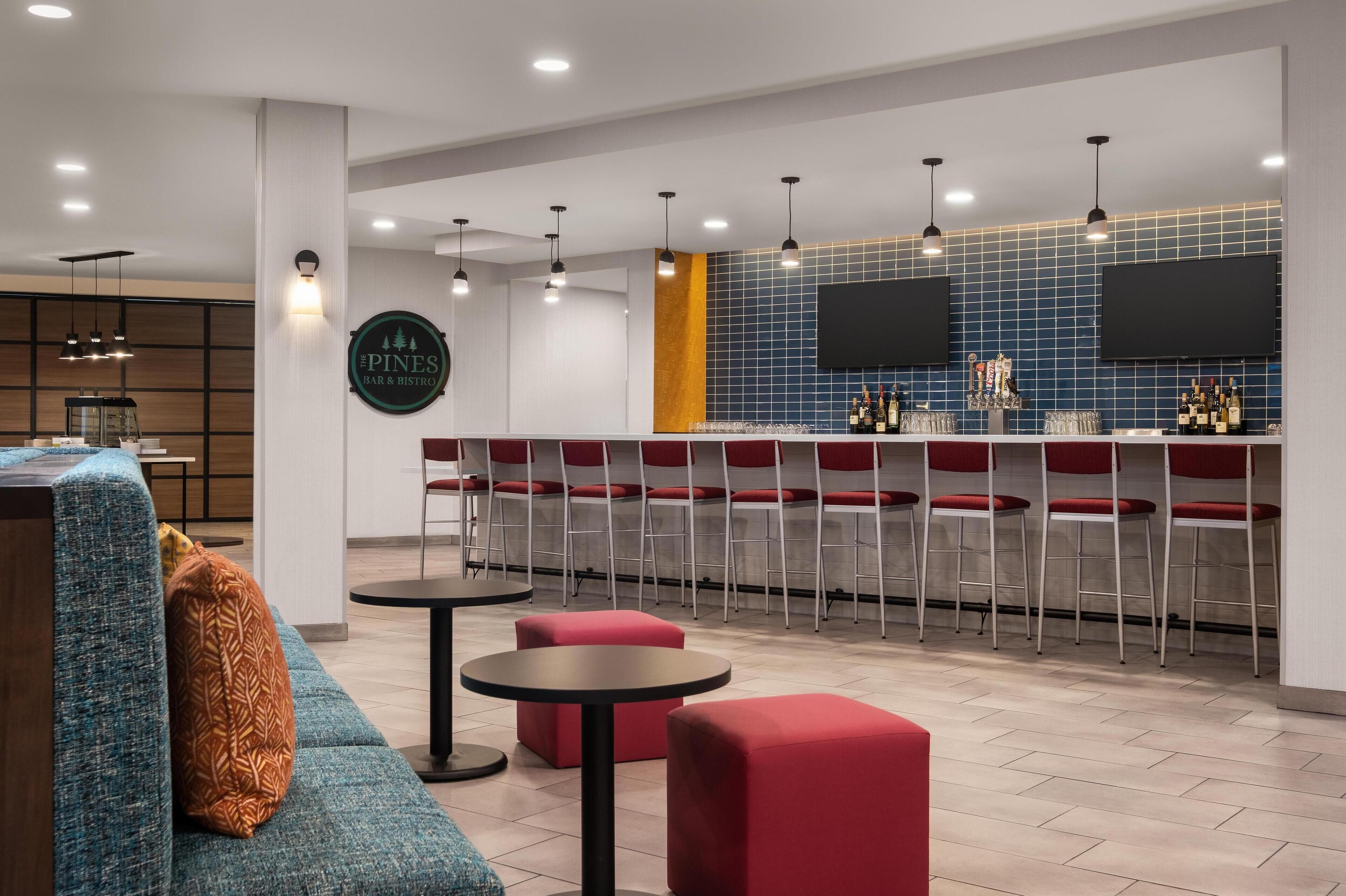 Un espace bar moderne avec des tabourets rouges, un comptoir doté d’un téléviseur et un coin salon avec un canapé bleu et des poufs rouges.