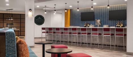 Un espace bar moderne avec des tabourets rouges, un comptoir doté d’un téléviseur et un coin salon avec un canapé bleu et des poufs rouges.